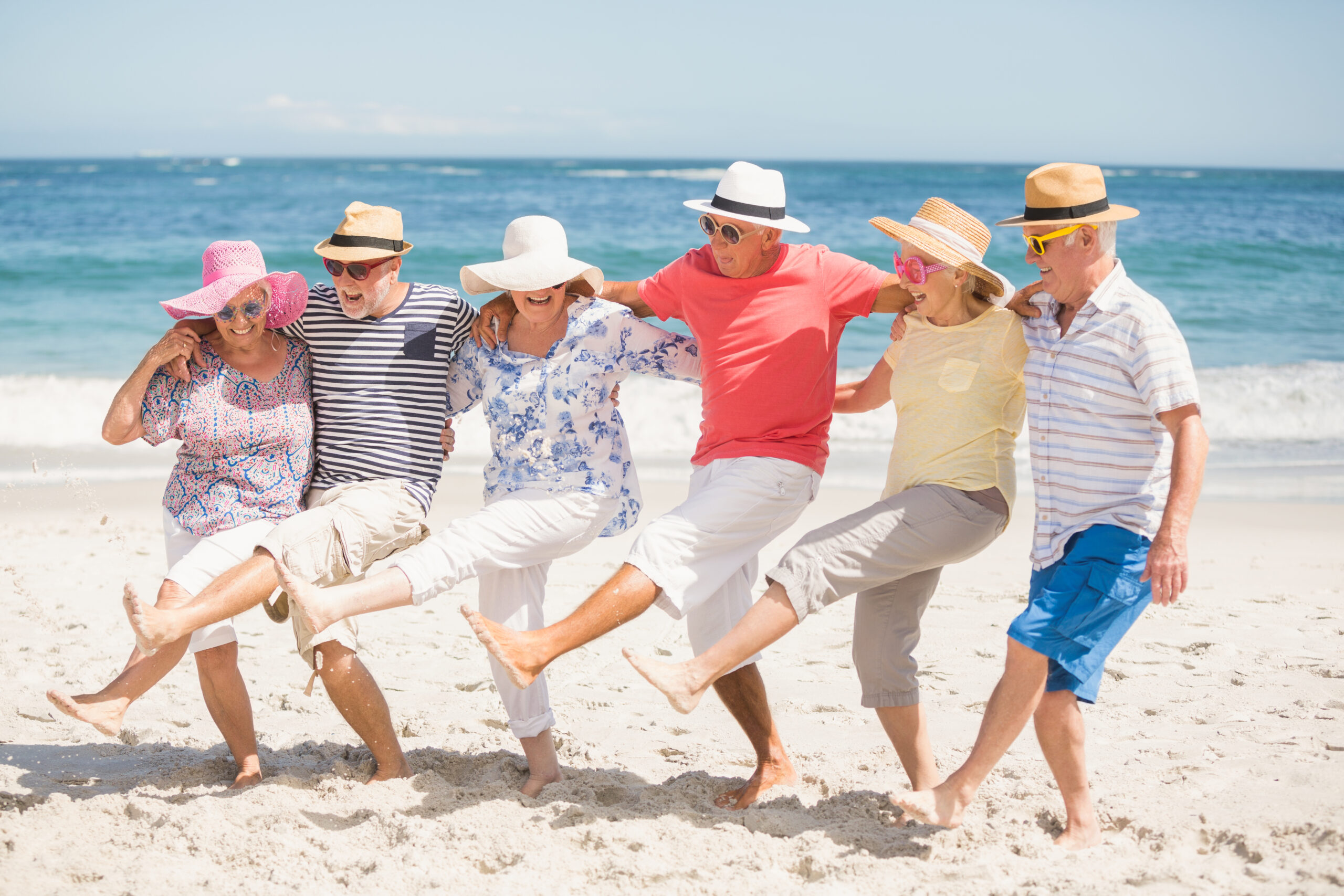 Group of older adults in hats, arms embraced, kicking happily at the beach